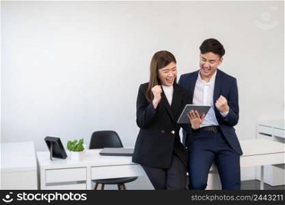 Successful young smart Asian businessman and businesswoman smiling and raising fist while looking achieved results online report on tablet. Business people wear formal suit working together at office