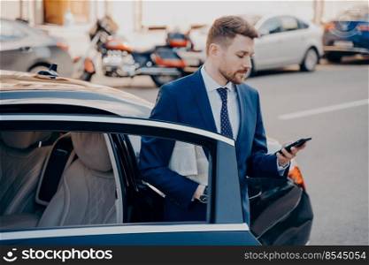 Successful young well groomed executive manager in formal stylish suit getting out of premium class car, arrival to workplace, while checking missed calls in his cell phone. Business people outdoors. Successful executive manager in formal stylish tuxedo suit getting out of car