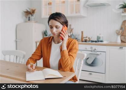 Successful young woman is call center assistant working from home. Spanish lady in orange shirt is remote employee. Girl is sitting at table at kitchen and taking notes into notebook.. Successful young woman is remote employee. Girl is sitting at table at kitchen and taking notes.