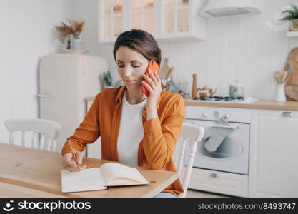 Successful young woman is call center assistant working from home. Spanish lady in orange shirt is remote employee. Girl is sitting at table at kitchen and taking notes into notebook.. Successful young woman is remote employee. Girl is sitting at table at kitchen and taking notes.