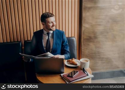 Succsessful smiling business man in blue formal suit looking outside window, holding newspaper while sitting in cafe with laptop computer after ordering coffee with croissant. Business man looking outside window in cafe while holding newspaper