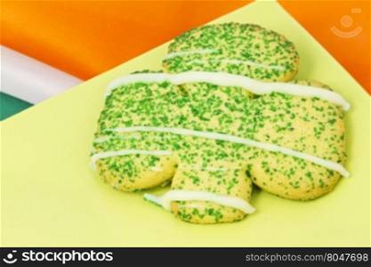 Sugar cookie cut in shamrock shape on green plate with orange, white, and green stripes of Irish flag visible as background. Close up with sugar sprinkles and frosting visible.