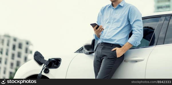 Suit-clad progressive businessman look at EV car&rsquo;s battery status from his phone while standing on a charging station with a power cable plug and a renewable energy-powered electric vehicle.. Suit-clad progressive businessman look at EV car&rsquo;s battery status from his phone