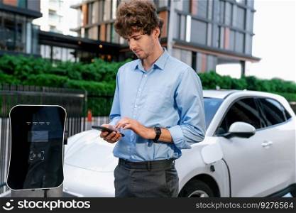 Suit-clad progressive businessman look at EV car&rsquo;s battery status from his phone while standing on a charging station with a power cable plug and a renewable energy-powered electric vehicle.. Suit-clad progressive businessman look at EV car&rsquo;s battery status from his phone