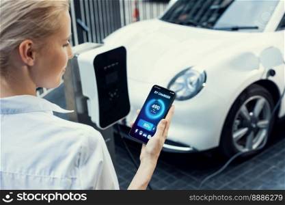 Suit-clad progressive businesswoman look at EV car’s battery status from her phone while standing on a charging station with a power cable plug and a renewable energy-powered electric vehicle.. Suit-clad progressive businesswoman look at EV car’s battery status from phone.