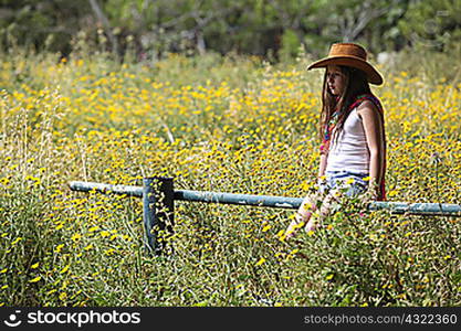 Sullen girl in cowboy hat sitting on fence in field