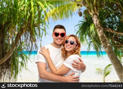 summer accessory, valentine&rsquo;s day and love concept - portrait of happy couple in white t-shirts and heart shaped sunglasses over tropical beach background in french polynesia. happy couple in white t-shirts and sunglasses