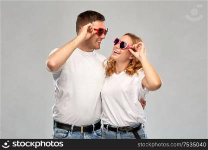 summer accessory, valentines day and love concept - portrait of happy couple in white t-shirts and heart shaped sunglasses over grey background. happy couple in white t-shirts and sunglasses