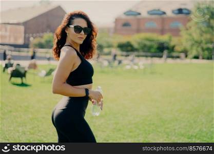 Summer activity and energy concept. Brunette sporty woman in sports clothes holds bottle of water wears sunglasses poses outdoor poses against blurred background with green lawn. Sporty lifestyle