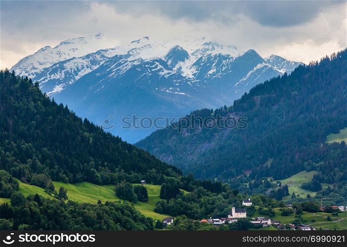 Summer Alps mountain landscape with village, fir forest on slope and snow covered rocky tops in far, Austria.