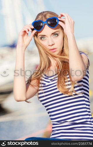 Summer and people concept. Fashion lovely blonde girl with heart shaped sunglasses in marina against yachts in port