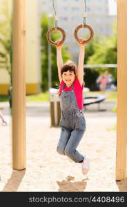 summer, childhood, leisure and people concept - happy little girl hanging on gymnastic rings at children playground