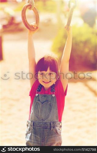 summer, childhood, leisure and people concept - happy little girl hanging on gymnastic rings at children playground. happy little girl on children playground