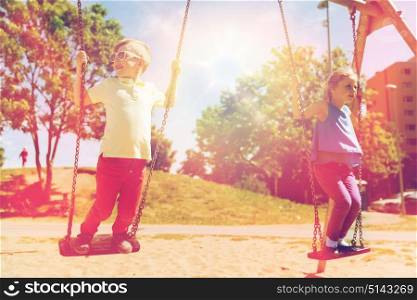 summer, childhood, leisure, friendship and people concept - two happy kids swinging on swing at children playground. two happy kids swinging on swing at playground