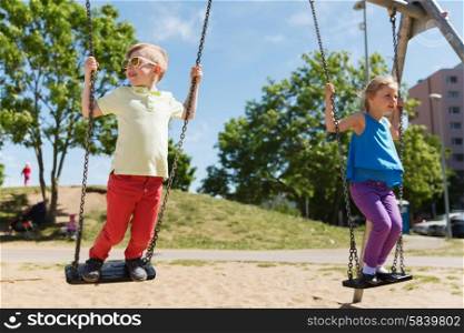 summer, childhood, leisure, friendship and people concept - two happy kids swinging on swing at children playground
