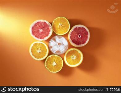 Summer cocktail ingredients with citrus fruits and glass with ice cubes on orange background in sunlight. Fresh oranges and grapefruit cut in half.