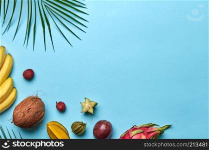 Summer composition of ripe sweet exotic fruits and palm branches on a blue background with space for text. Flat lay. Corner frame of a green palm branch and a variety of tropical fruits on a blue background with space for text. Flat lay