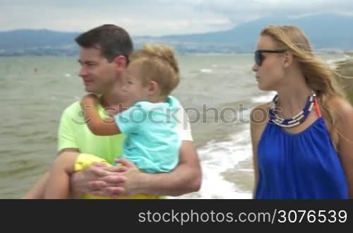 Summer day, a young family is walking along the beach, having a lovely conversation and smiling. Dad carries the baby, mother is going near them. They enjoy a warm summer day and each other&acute;s company