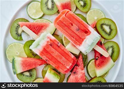 Summer dessert from frozen fruit juice lolly with ice and pieces of watermelon, kiwi and lime in a plate on a gray marble background. Flat lay. Frozen watermelon smoothies on a stick in a bowl with pieces of kiwi, lime, watermelon and slices of ice on a gray marble background. Flat lay