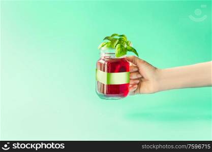 Summer drink in female hand. Red refreshing berries beverage in glass jar with green white branding mock up at sunny bight mint background. Summer mood. Blank Label at juice bottle