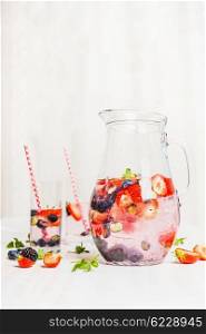 Summer drink with berries in jug and glasses with straw on white wooden background, side view, vertical