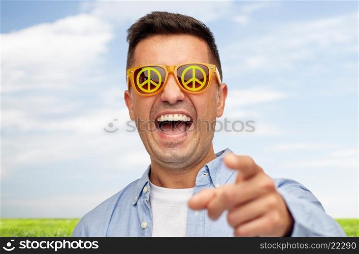 summer, ecology, emotions and people concept - face of laughing middle aged man in sunglasses with green peace symbol pointing finger on you over blue sky and grass background
