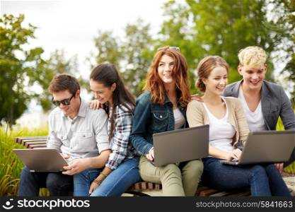 summer, education, technology and people concept - group of students or teenagers with laptop computers sitting on bench outdoors
