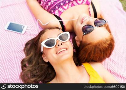 summer fashion, leisure and technology concept - smiling teenage girls in sunglasses and earphones lying on picnic blanket and listening to music from smartphone. teenage girls listening to music from smartphone