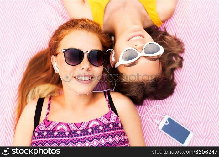 summer fashion, leisure and technology concept - smiling teenage girls in sunglasses and earphones lying on picnic blanket and listening to music from smartphone. teenage girls listening to music from smartphone