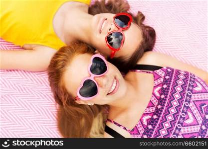 summer fashion, leisure and valentines day concept - smiling teenage girls in heart shaped sunglasses lying on picnic blanket. teenage girls in sunglasses on picnic blanket