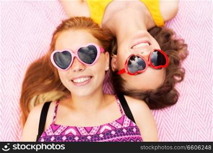summer fashion, leisure and valentines day concept - smiling teenage girls in heart shaped sunglasses lying on picnic blanket. teenage girls in sunglasses on picnic blanket