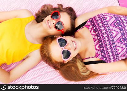 summer fashion, leisure and valentines day concept - smiling teenage girls in heart shaped sunglasses lying on picnic blanket. teenage girls in sunglasses on picnic blanket