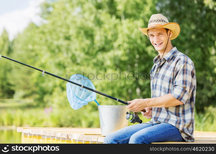 Summer fishing. Young guy in hat sitting on bridge and fishing