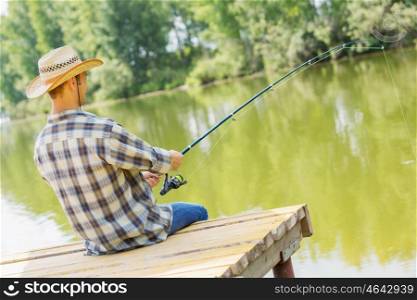 Summer fishing. Young guy in hat sitting on bridge and fishing