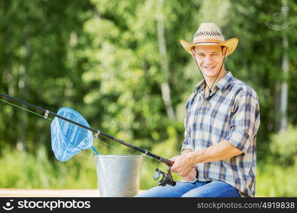 Summer fishing. Young guy in hat sitting on bridge and fishing