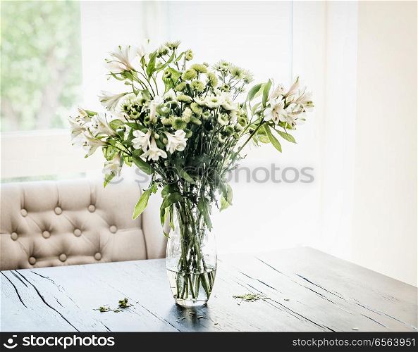 Summer flowers bunch in vase on table at window in living room with falling petals. Cozy home and house decoration