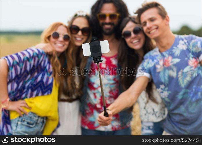 summer holidayds, technology and people concept - smiling young hippie friends in sunglasses taking picture by smartphone on selfie stick in countryside. hippie friends with smartphone and selfie stick