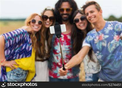 summer holidayds, technology and people concept - smiling young hippie friends in sunglasses taking picture by smartphone on selfie stick in countryside. hippie friends with smartphone and selfie stick