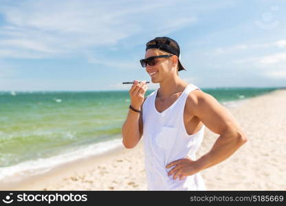 summer holidays and people concept - happy smiling young man in sunglasses using voice command recorder on smartphone on beach. smiling man calling on smartphone on summer beach