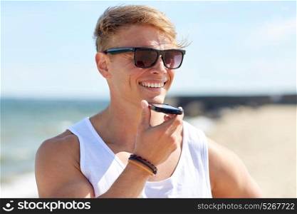summer holidays and people concept - happy smiling young man in sunglasses using voice command recorder on smartphone on beach. smiling man calling on smartphone on summer beach