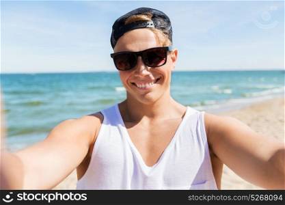 summer holidays and people concept - happy smiling young man in sunglasses and hat taking selfie on beach. man in sunglasses taking selfie on summer beach