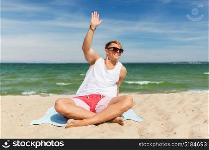 summer holidays and people concept - happy smiling young man sunbathing and greeting someone on beach towel. happy smiling young man sunbathing on beach towel