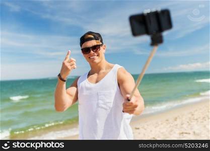 summer holidays and people concept - happy smiling young man with smartphone selfie stick taking picture on beach. man with smartphone selfie stick on summer beach