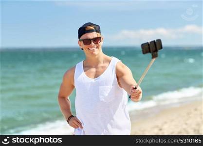summer holidays and people concept - happy smiling young man with smartphone selfie stick taking picture on beach. man with smartphone selfie stick on summer beach