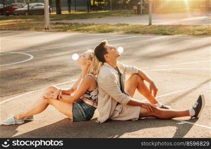 summer holidays and people concept - happy young couple sitting back to back and blowing bubble gums on basketball playground. couple sitting on ground and blowing bubble gums