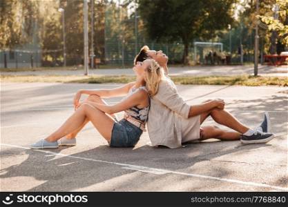 summer holidays and people concept - happy young couple sitting back to back on basketball playground. happy couple sitting on basketball playground