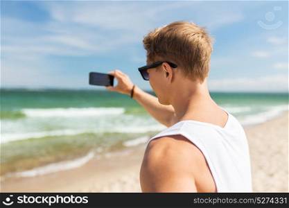 summer holidays and people concept - young man with smartphone on beach photographing sea. man with smartphone photographing on summer beach