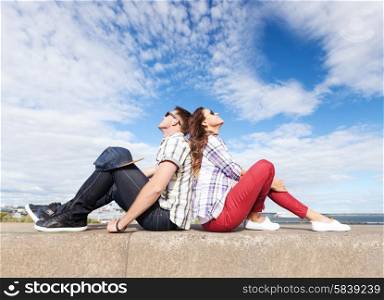 summer holidays and teenage concept - teenagers sitting back to back and looking up in the sky. teenagers sitting back to back