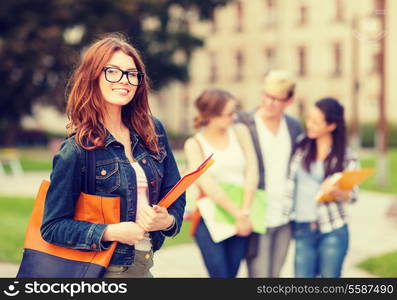 summer holidays, education, campus and teenage concept - smiling female student in black eyeglasses with folders and group in the back