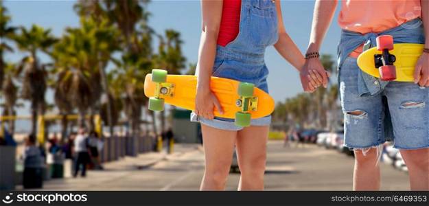 summer holidays, extreme sport and relationships concept - close up of happy teenage couple with short modern cruiser skateboards holding hands over venice beach in los angeles background. couple with skateboards at venice beach in la